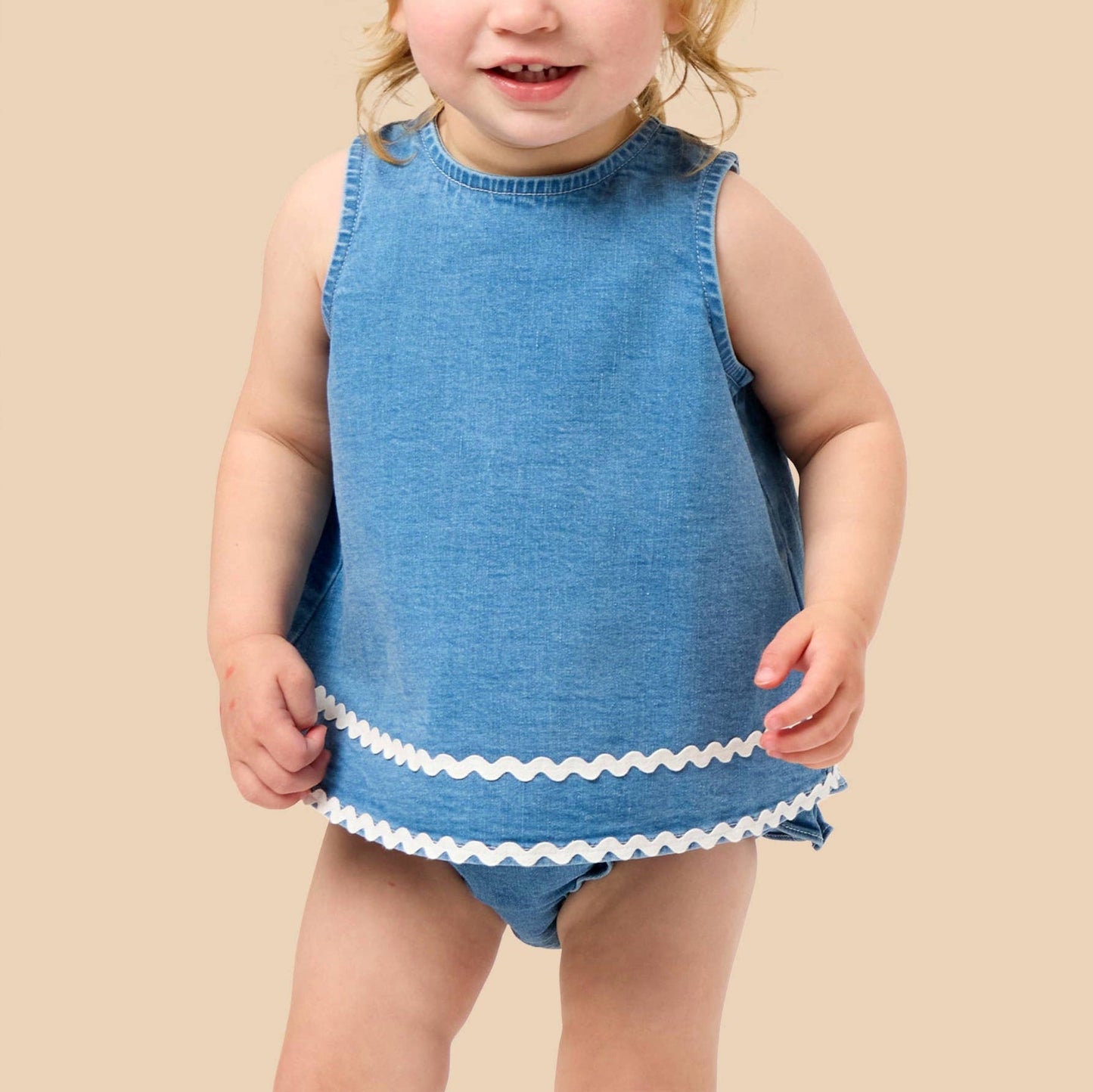 A young toddler girl with blonde hair wearing a sleeveless blue denim-like romper with white scalloped trim at the hem, standing against a solid beige background.