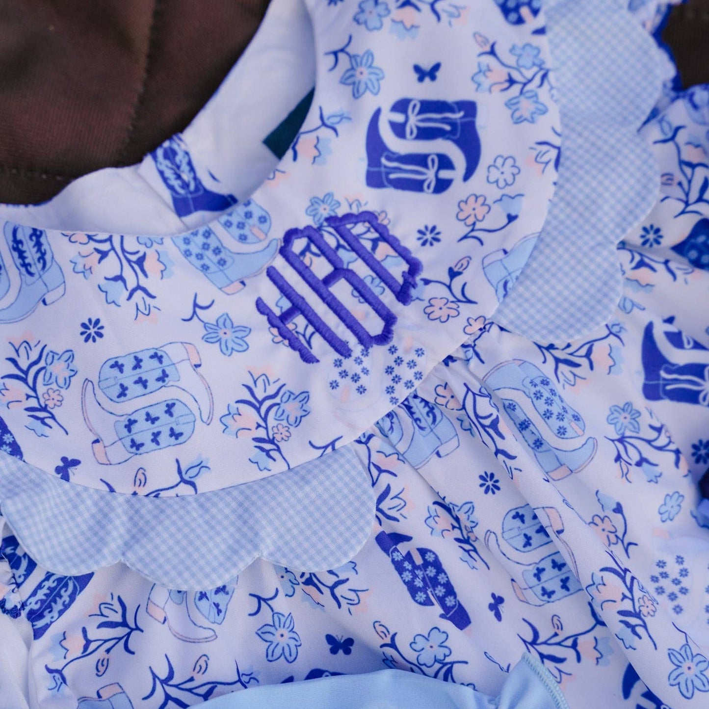 Close-up of a baby outfit featuring a white and blue patterned dress with a ruffled collar, blue monogram embroidery, and light blue ruffled trim.