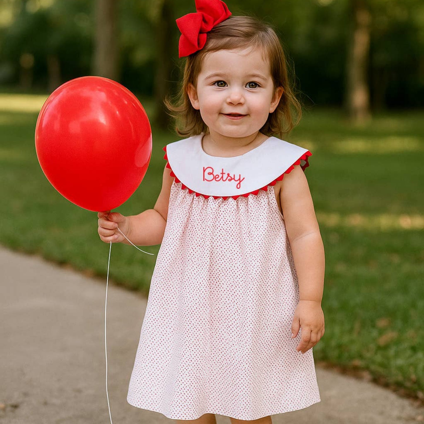 Child in a white dress with red trim holding a red balloon outdoors.