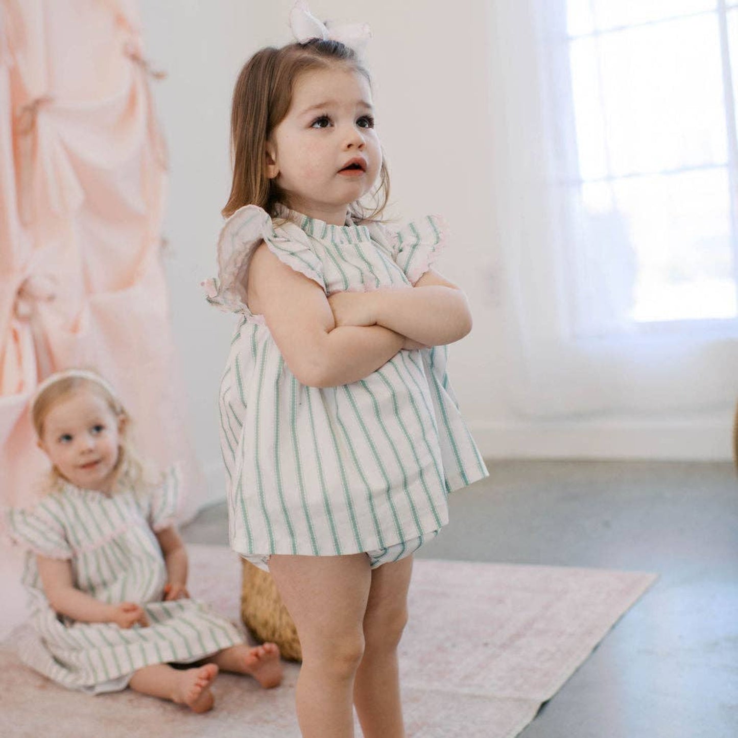 Young girl in a striped dress standing on a light-colored rug with a window in the background.