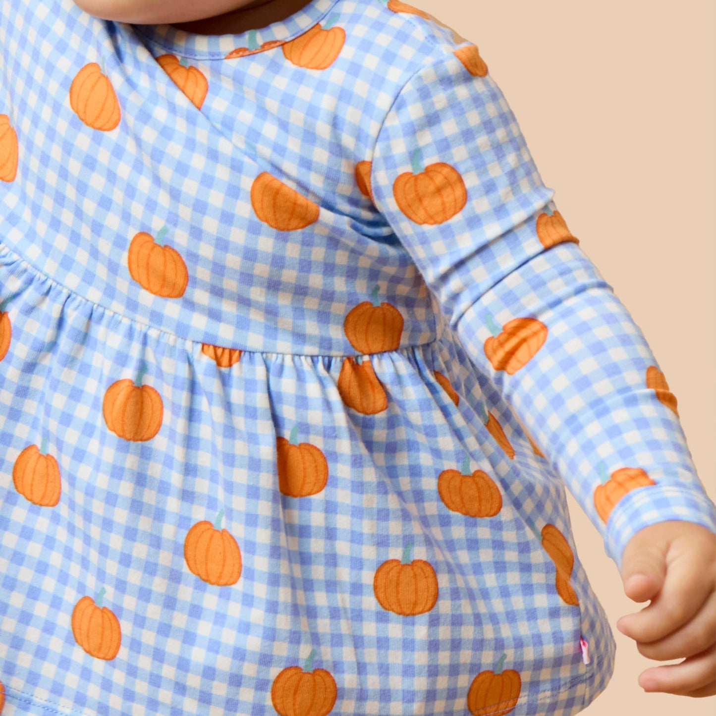 A close-up of a baby wearing a light blue and white gingham long-sleeve dress with an orange pumpkin print, set against a solid beige background.