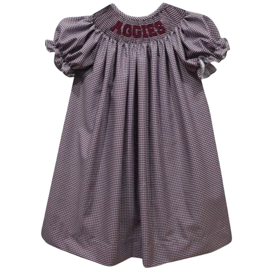 A toddler girl's smocked dress with a small purple and white gingham pattern, featuring puff sleeves and 'AGGIES' embroidered in maroon on the back neckline.