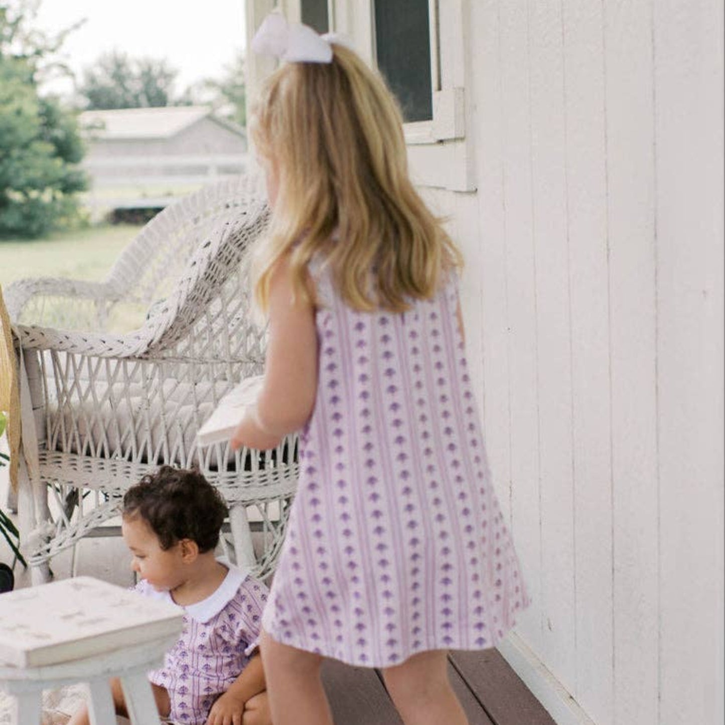 A young girl with blonde hair in a white bow stands on a wooden porch wearing a lavender and white patterned sleeveless dress and white shoes. A smaller child sits on the porch nearby.