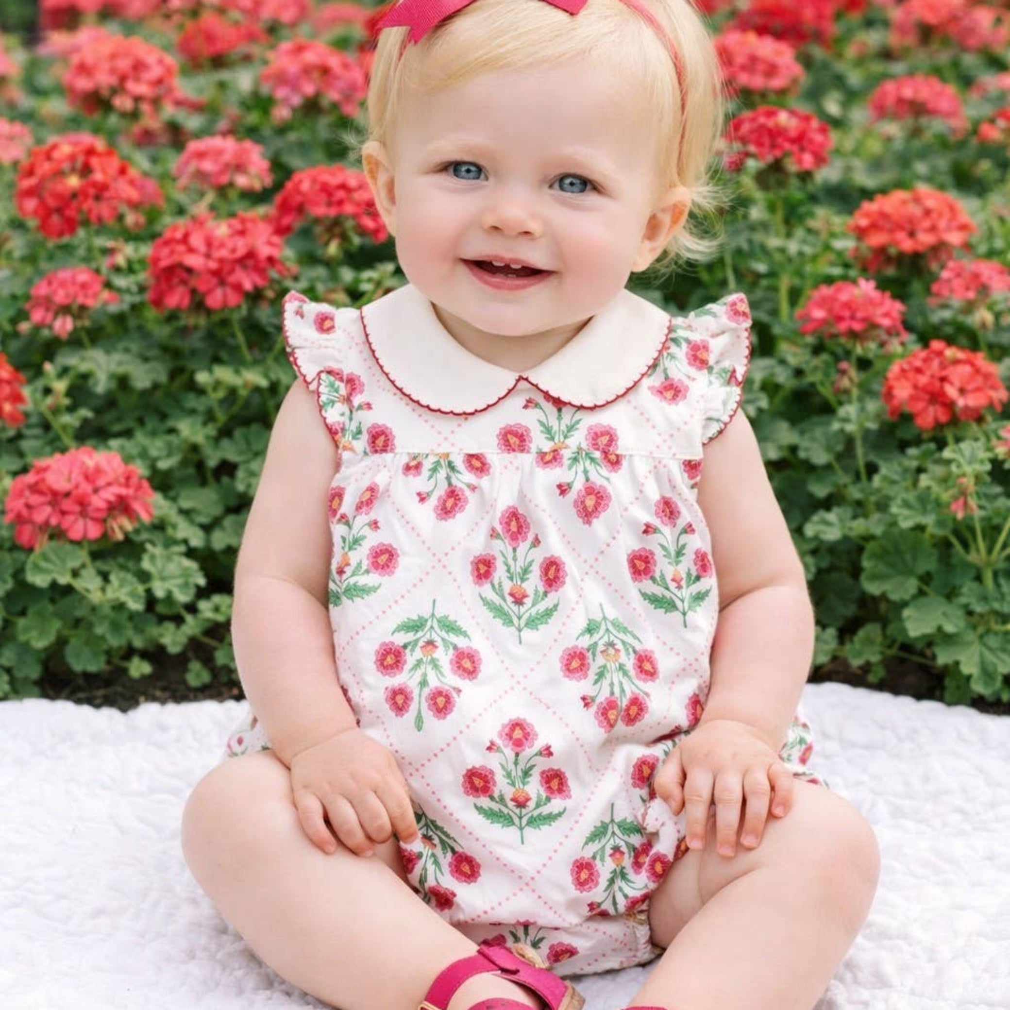 Child wearing a floral dress and pink sandals sitting in front of red flowers.