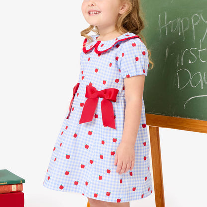 Young girl in a checkered dress with red polka dots standing next to a chalkboard with 'Happy First Day' written on it.