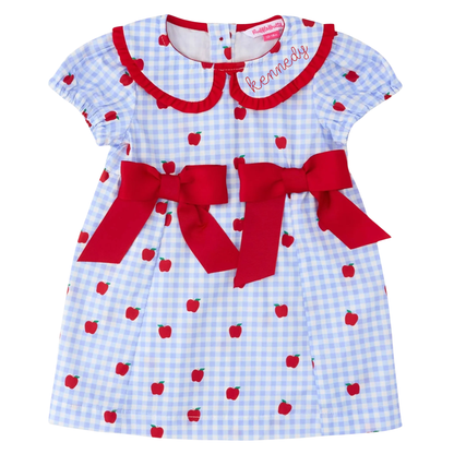 A baby girl's short-sleeve dress with a light blue and white gingham pattern, decorated with red apple prints, a red collar, and two large red bows at the waist. The name 'kennedy' is embroidered in red on the collar.