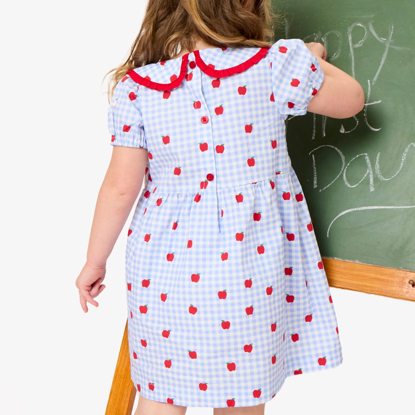 Child wearing a dress with red polka dots and a large red bow, standing in front of a chalkboard.