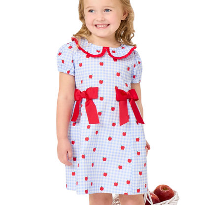 A young girl wearing a light blue and white gingham dress with red apple prints, red ruffled collar trim, and red ribbon bows at the waist, standing against a white background.