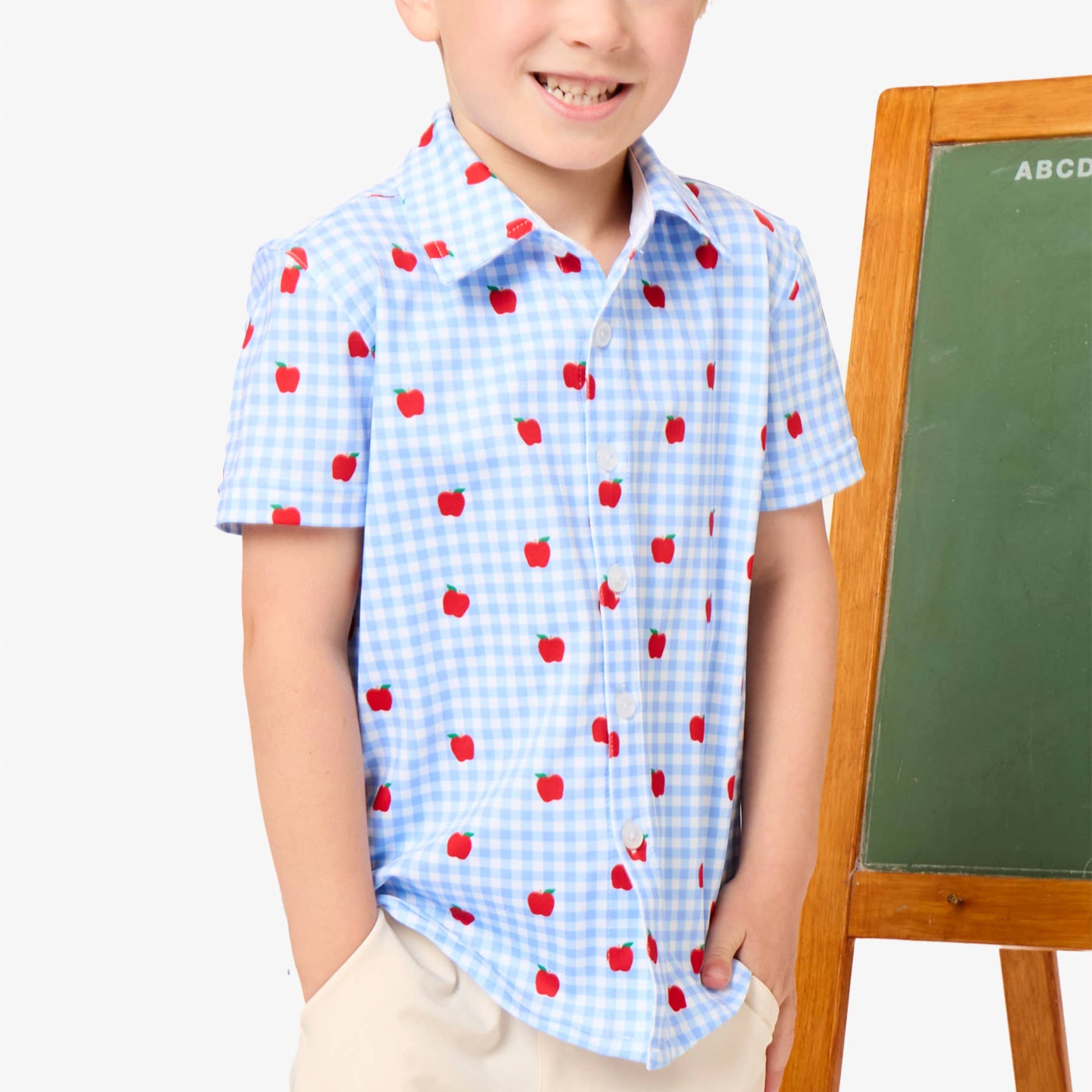 A young boy with blond hair stands next to a chalkboard, wearing a light blue and white gingham short-sleeve button-up shirt with a red apple print, paired with beige pants.