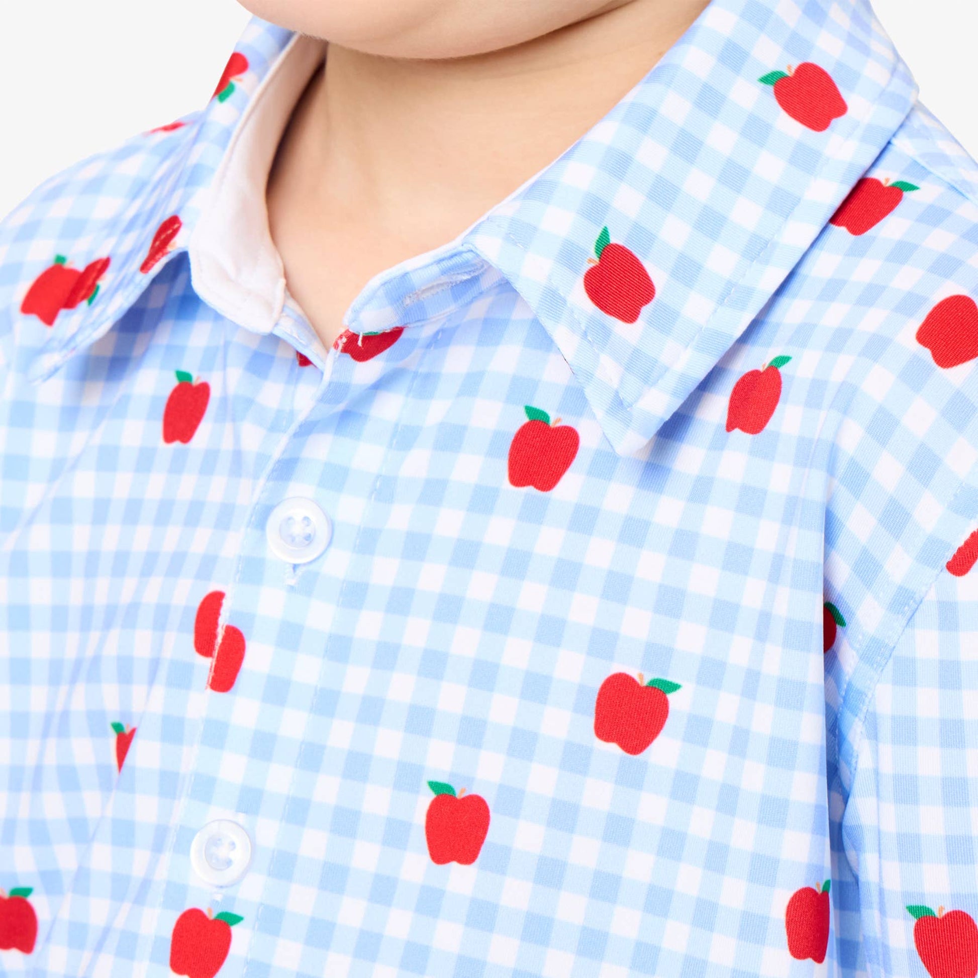 A close-up of a child wearing a light blue and white gingham short-sleeve button-up shirt with a red apple print.
