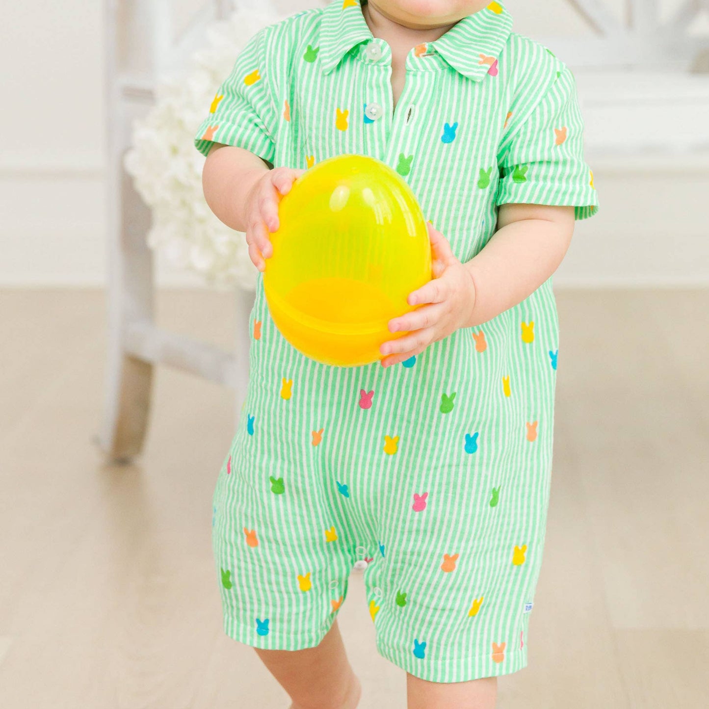 Baby in a green outfit with colorful patterns holding a yellow toy.