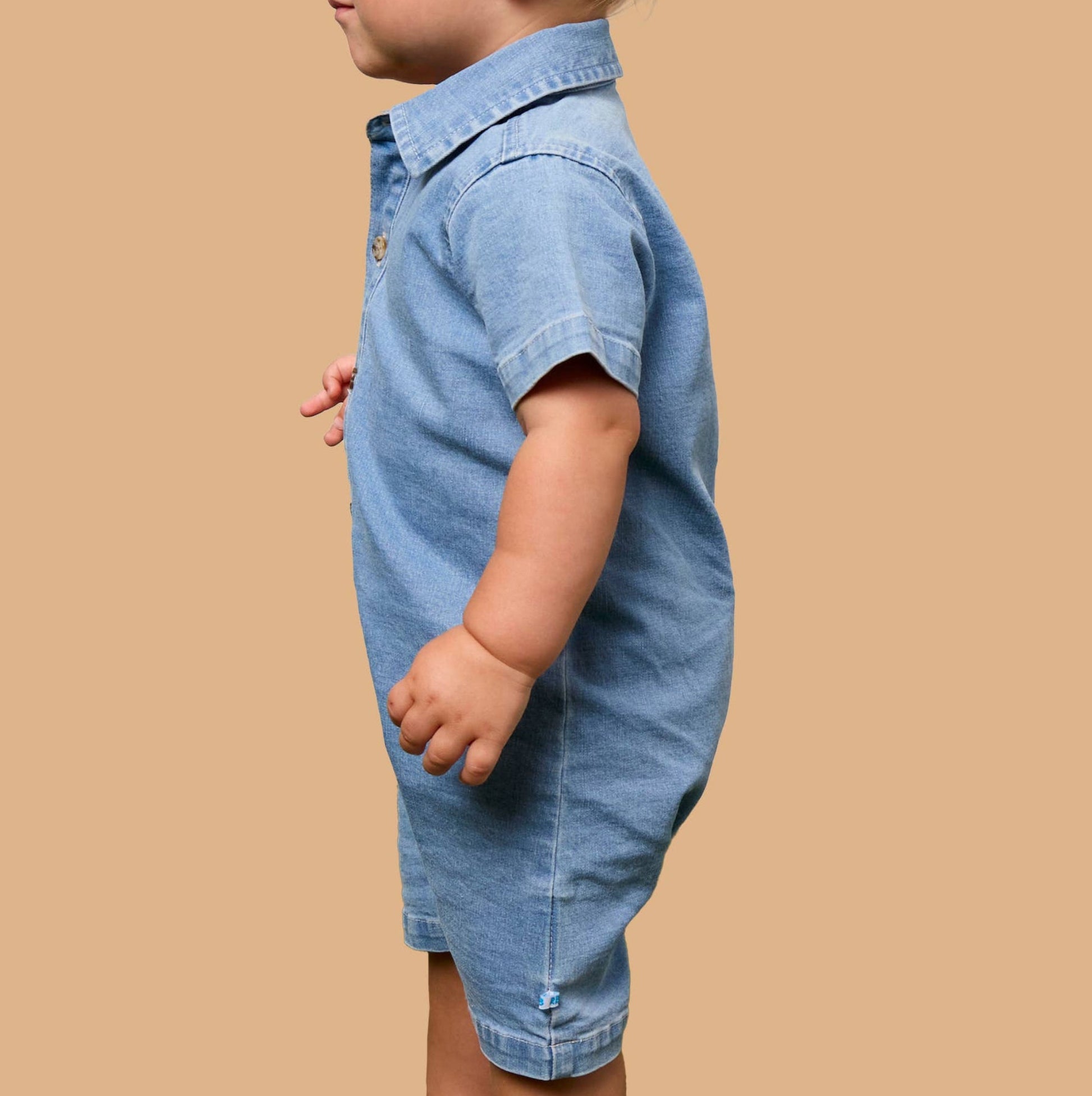 A toddler boy in profile wearing a light blue denim short-sleeve romper, standing against a solid tan background.