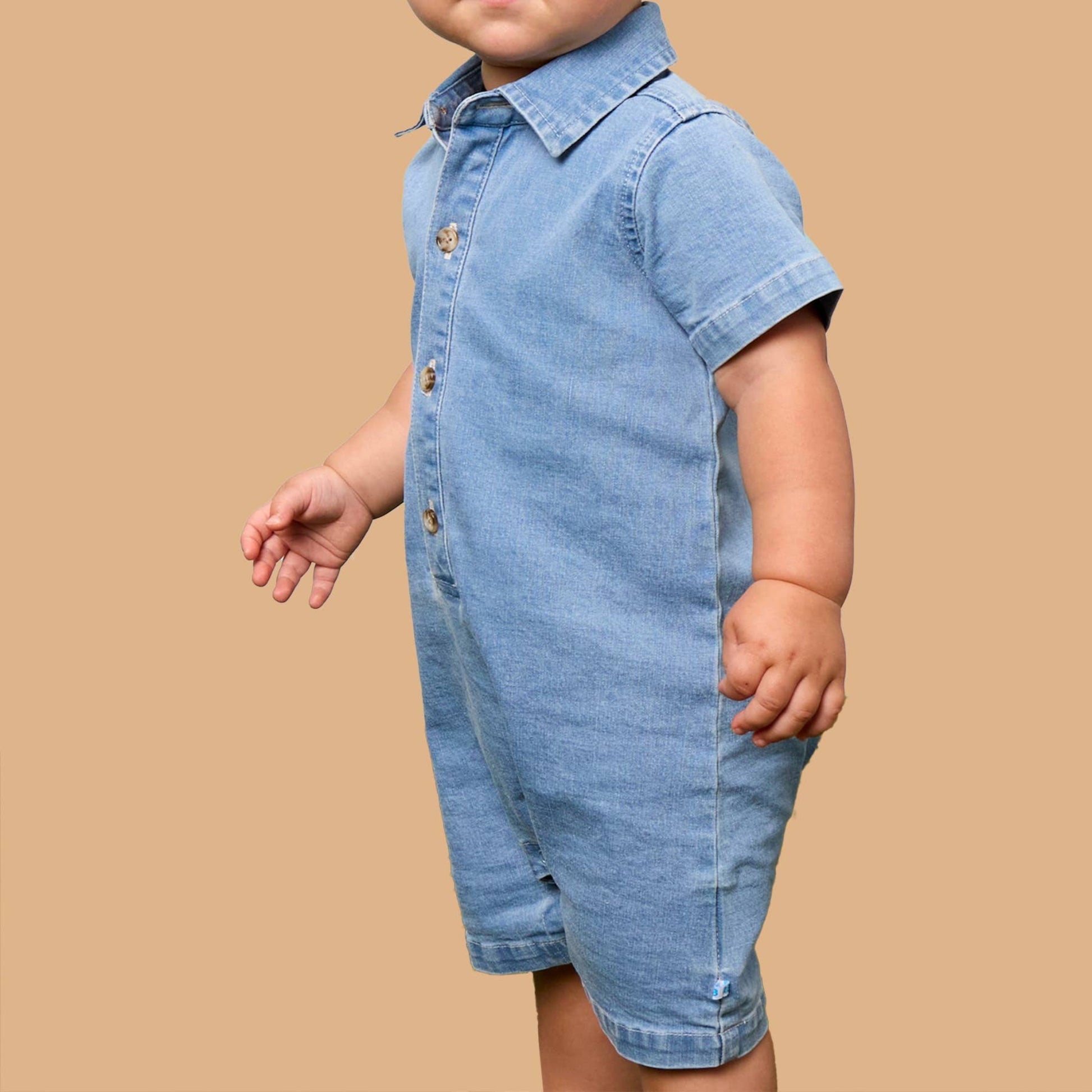 A toddler boy with blond hair wearing a light blue denim short-sleeve romper, standing against a solid tan background.