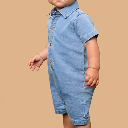 A toddler boy with blond hair wearing a light blue denim short-sleeve romper, standing against a solid tan background.