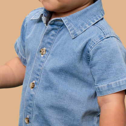 A close-up of a young child wearing a light blue denim short-sleeve button-up shirt against a solid tan background.