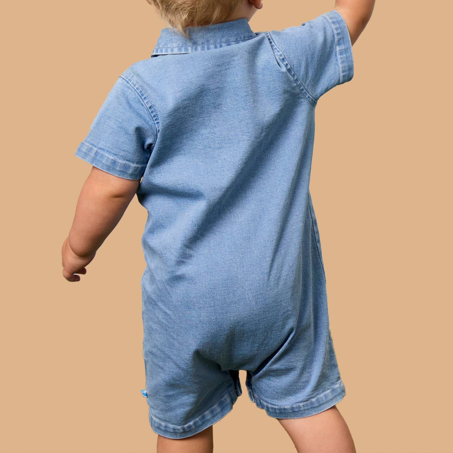 A toddler viewed from behind wearing a light blue denim short-sleeve romper, standing against a solid tan background.