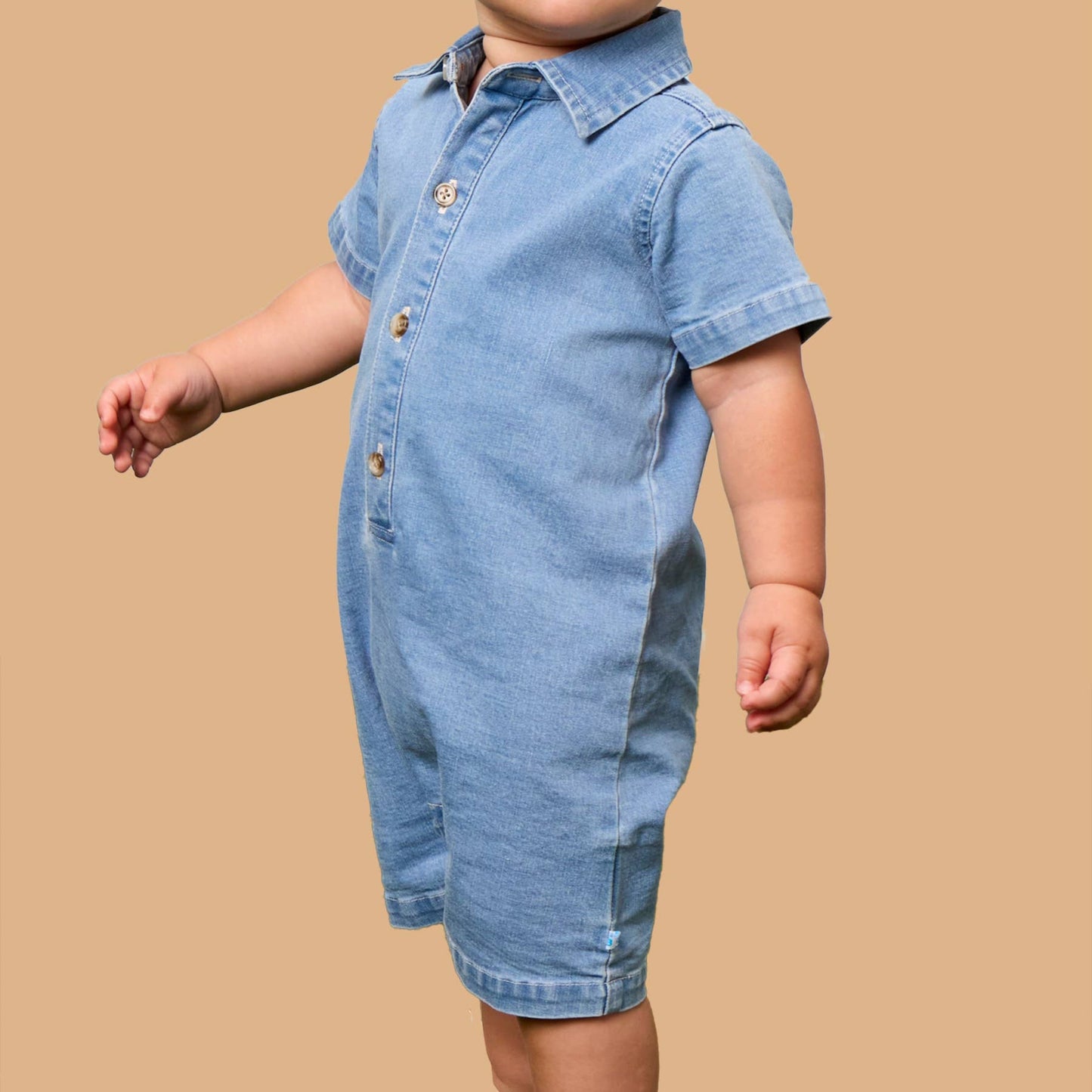 A toddler standing against a solid tan background, wearing a light blue denim short-sleeve romper with a button-down front.