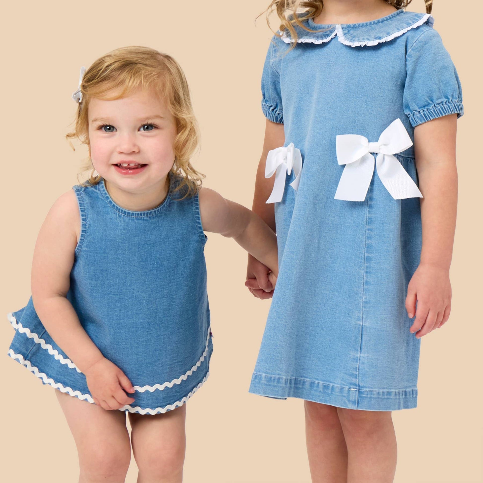 Two young girls wearing matching blue dresses with white ruffle trim and large white bows, standing against a solid beige background.