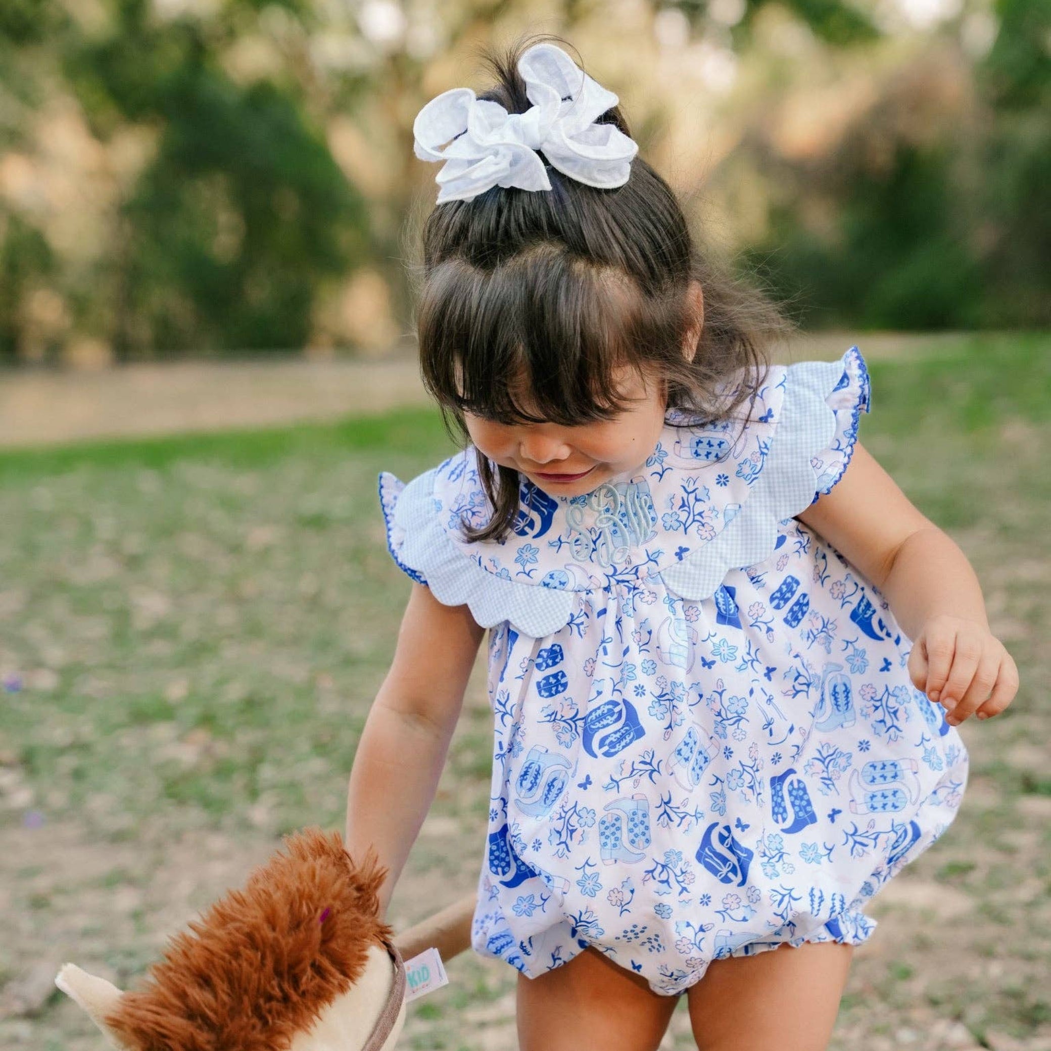 A toddler girl wearing a blue and white patterned romper with a white bow in her hair, holding a plush toy horse in an outdoor park setting.