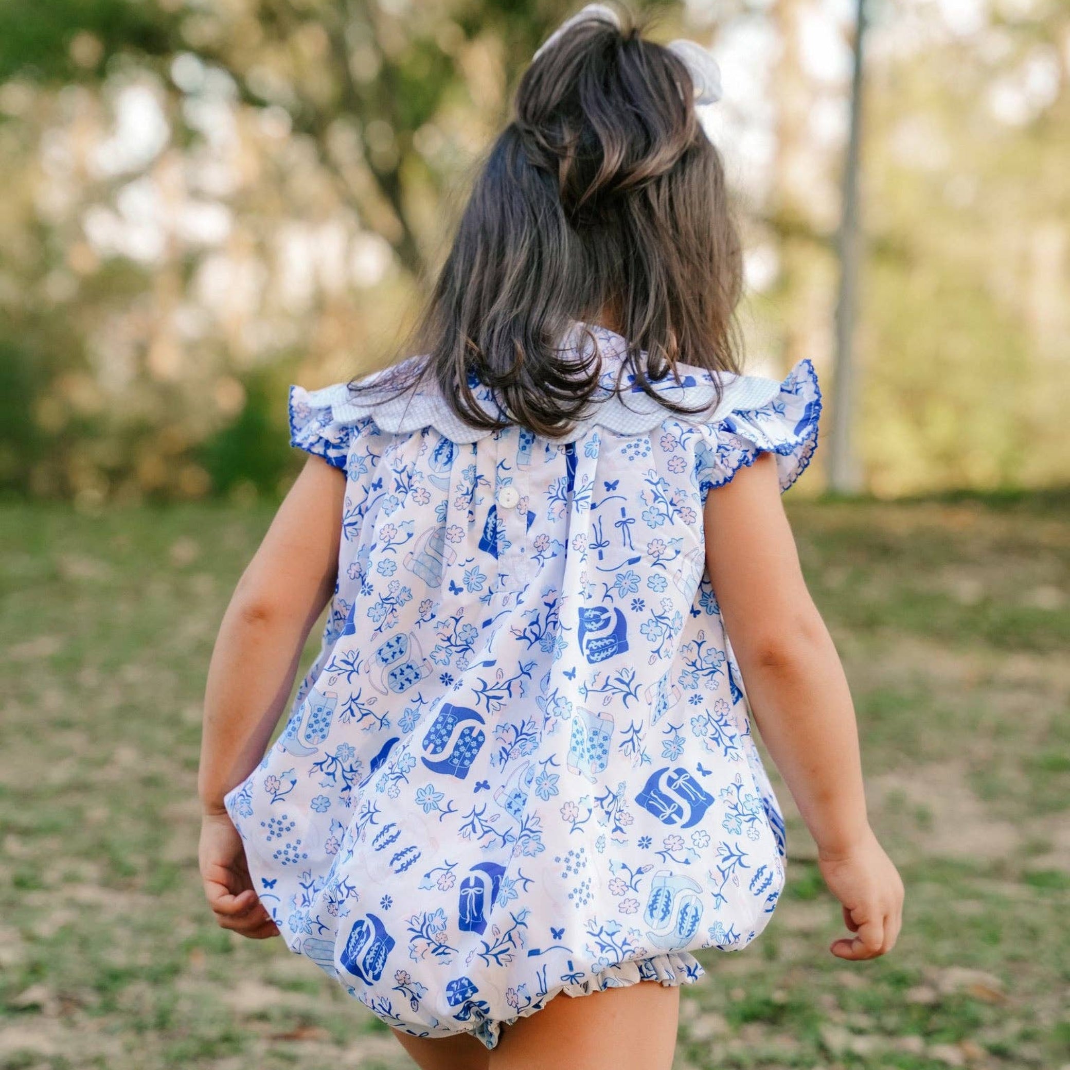 A young girl seen from behind wearing a white and blue patterned ruffle-sleeve romper, walking outdoors on a grassy path with trees in the background.