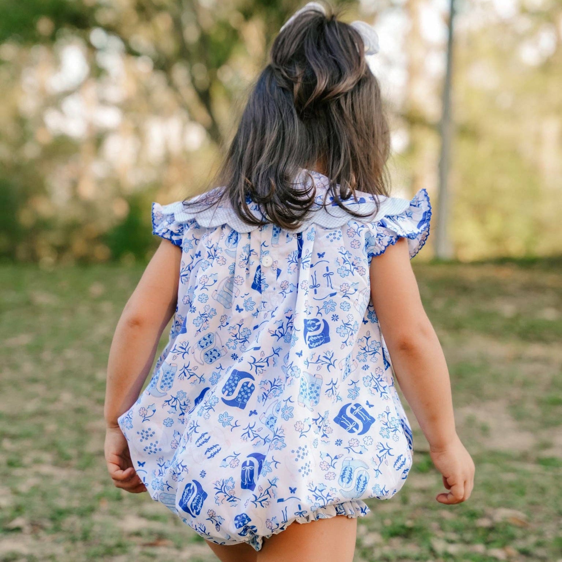 A young girl seen from behind wearing a white and blue patterned ruffle-sleeve romper, walking outdoors on a grassy path with trees in the background.