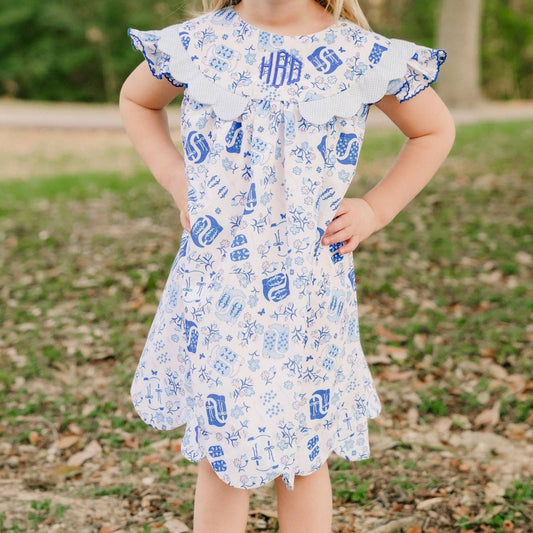 A young girl stands outdoors in a park-like setting wearing a white tiered dress with a blue monogram pattern, a matching blue bow in her hair, and cream cowboy boots.