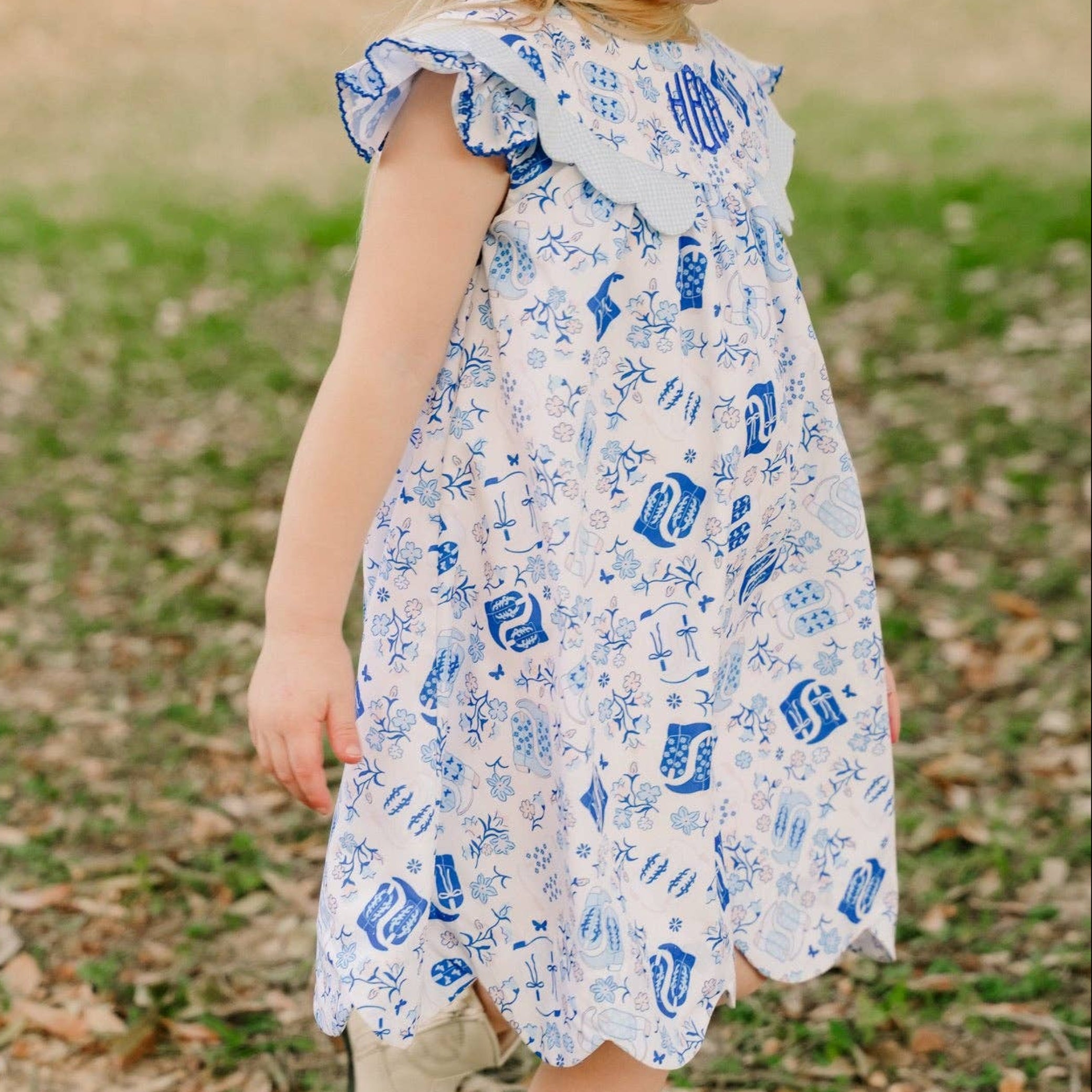 A young girl wearing a white dress with a blue nautical print and a blue bow in her hair, walking outdoors on a grassy path.