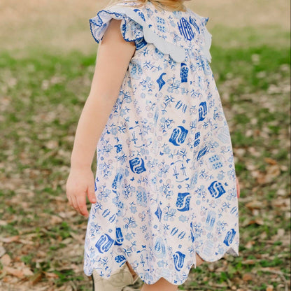 A young girl wearing a white dress with a blue nautical print and a blue bow in her hair, walking outdoors on a grassy path.