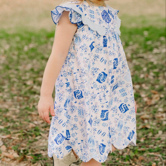A young girl wearing a white dress with a blue nautical print and a blue bow in her hair, walking outdoors on a grassy path.