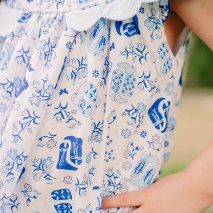 Close-up of a child wearing a sleeveless white dress with a blue floral and paisley print, featuring a ruffled tie at the neckline and a side pocket being touched by a hand.