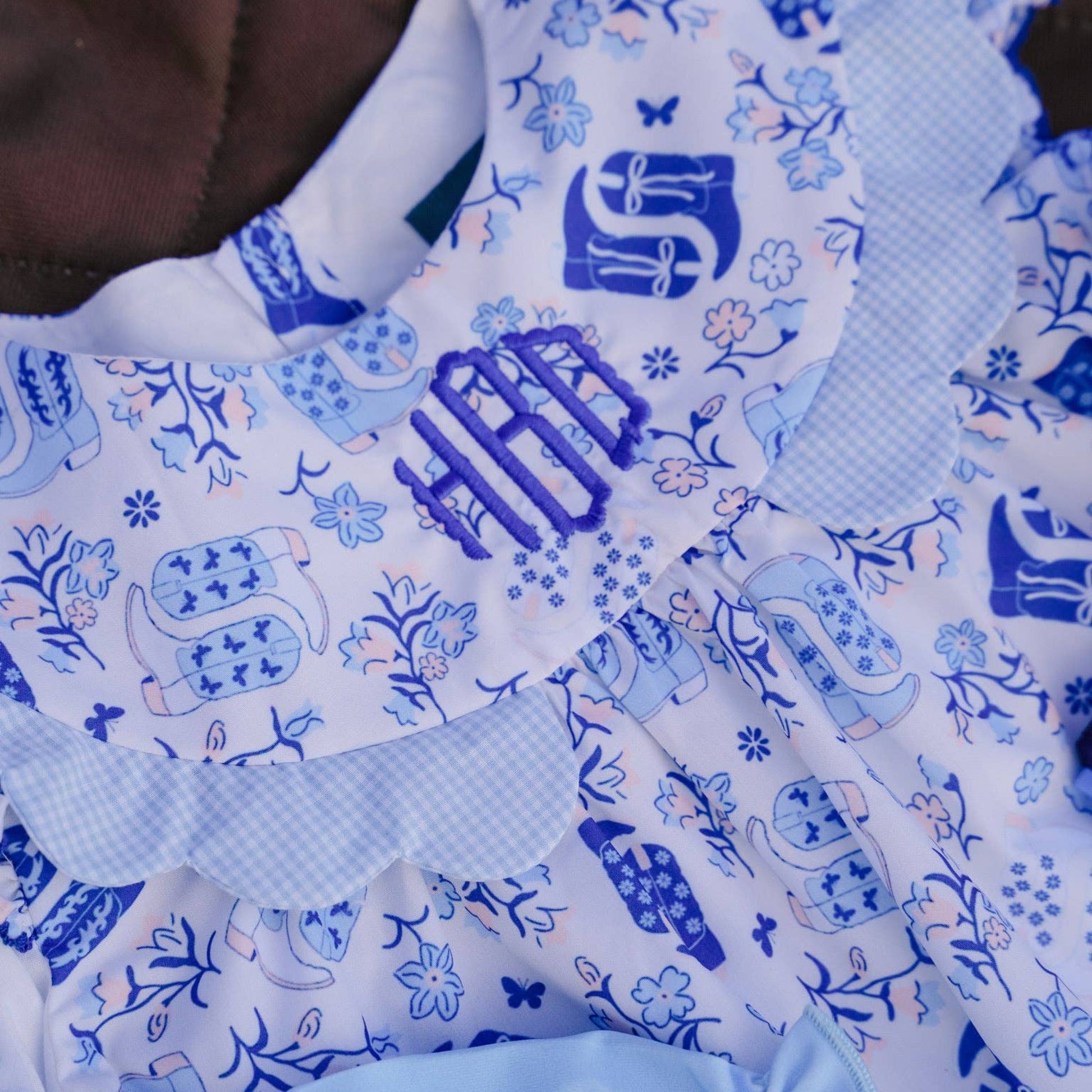Close-up of a baby outfit featuring a white and blue patterned dress with a ruffled collar, blue monogram embroidery, and light blue ruffled trim.