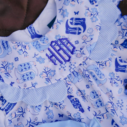 Close-up of a baby outfit featuring a white and blue patterned dress with a ruffled collar, blue monogram embroidery, and light blue ruffled trim.