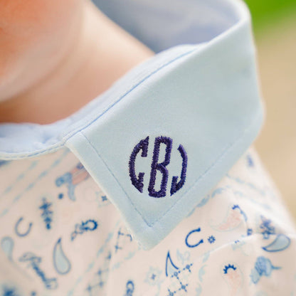 Close-up of a baby's neck and shoulder wearing a light blue collared shirt with a navy monogram 'CBJ' on the collar and a pale pink patterned shirt underneath.