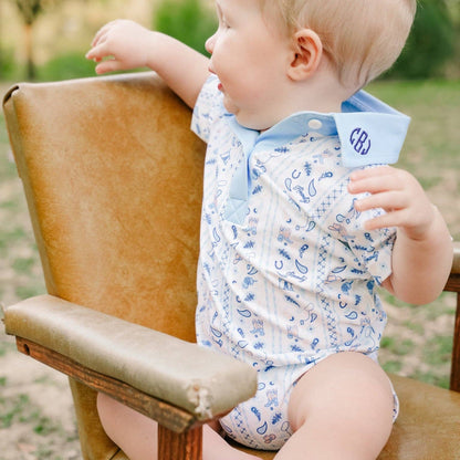A baby wearing a light blue and white paisley short-sleeve bodysuit sits on a tan leather chair outdoors, looking to the side.