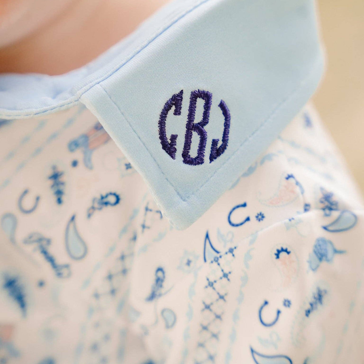 Close-up of a baby's neck and shoulder wearing a light blue collared shirt with a navy monogram 'CBJ' on the collar and a pale pink patterned shirt underneath.