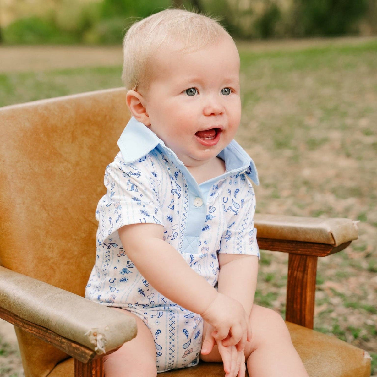 A baby sitting on a brown leather armchair outdoors, wearing a light blue and white patterned short-sleeve shirt and matching diaper cover.