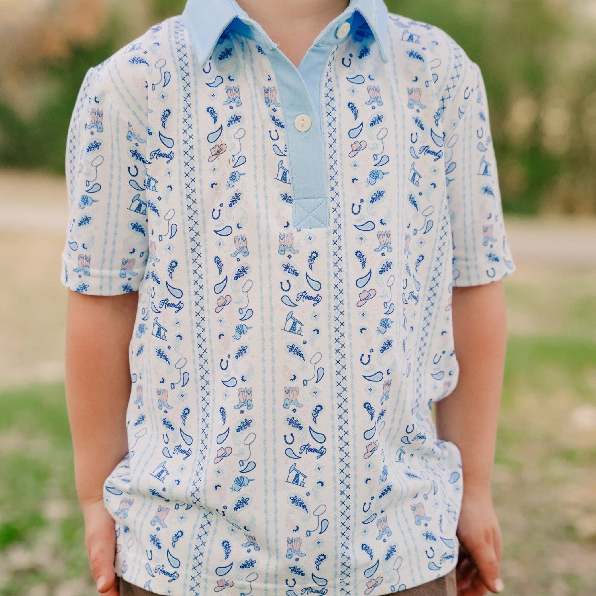 A young boy wearing a short-sleeve button-up shirt with vertical blue paisley and stripe patterns on a white base, paired with brown pants, standing outdoors with a blurred green background.