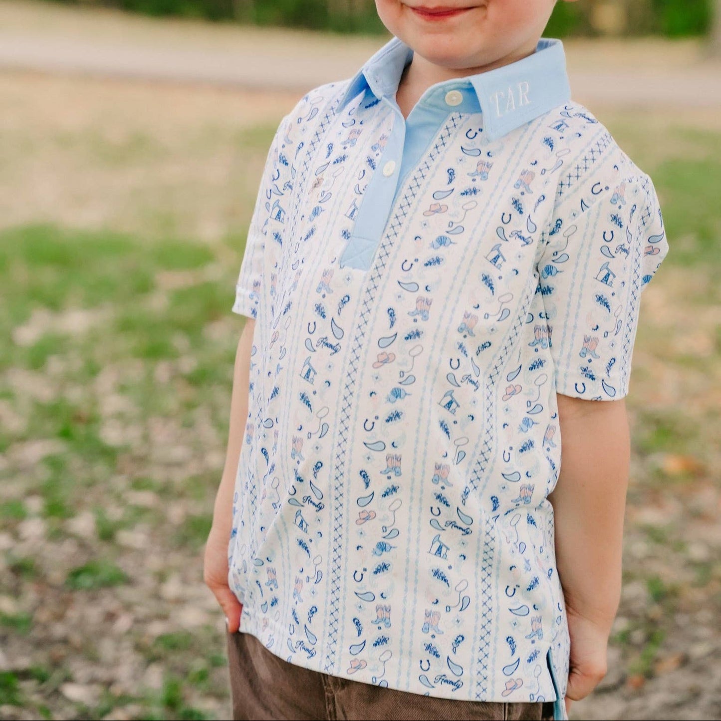 A young boy with short blond hair smiling outdoors, wearing a light blue and white patterned short-sleeve polo shirt.