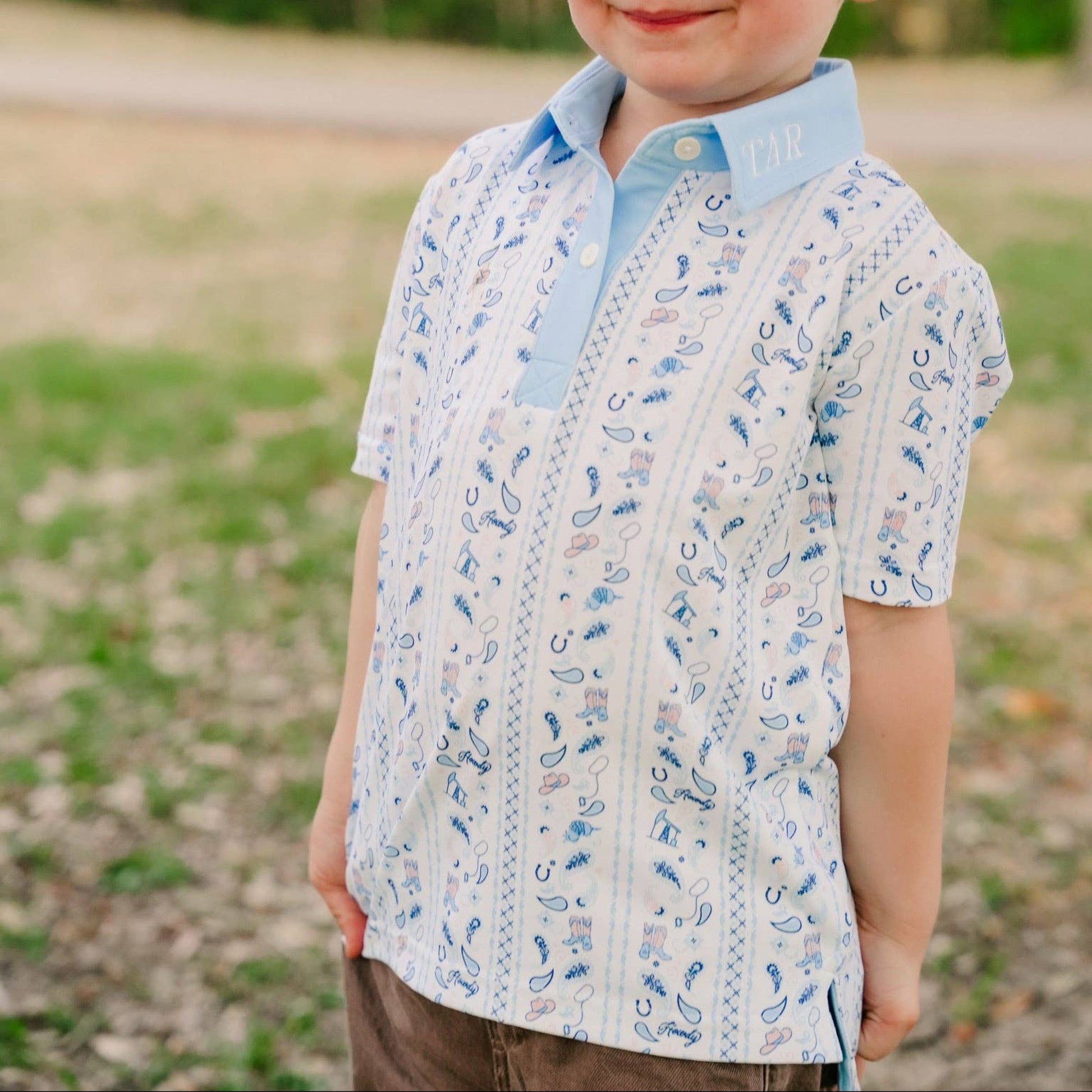 A young boy with short blond hair smiling outdoors, wearing a light blue and white patterned short-sleeve polo shirt.
