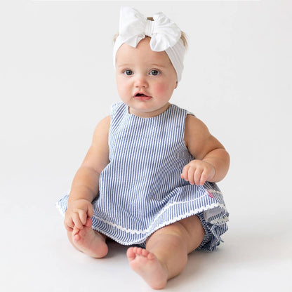 Baby wearing a blue and white seersucker dress with a white headband on a light background