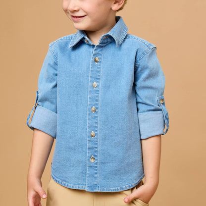 A young boy with blond hair wearing a light blue denim button-down shirt with rolled-up sleeves and tan pants, standing against a solid tan background.