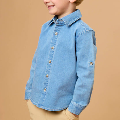 A young boy with blonde hair wearing a light blue denim long-sleeve button-up shirt and khaki pants, standing against a solid tan background.