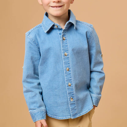A young boy with blond hair wearing a light blue denim long-sleeve button-up shirt and khaki pants, standing against a solid tan studio background.
