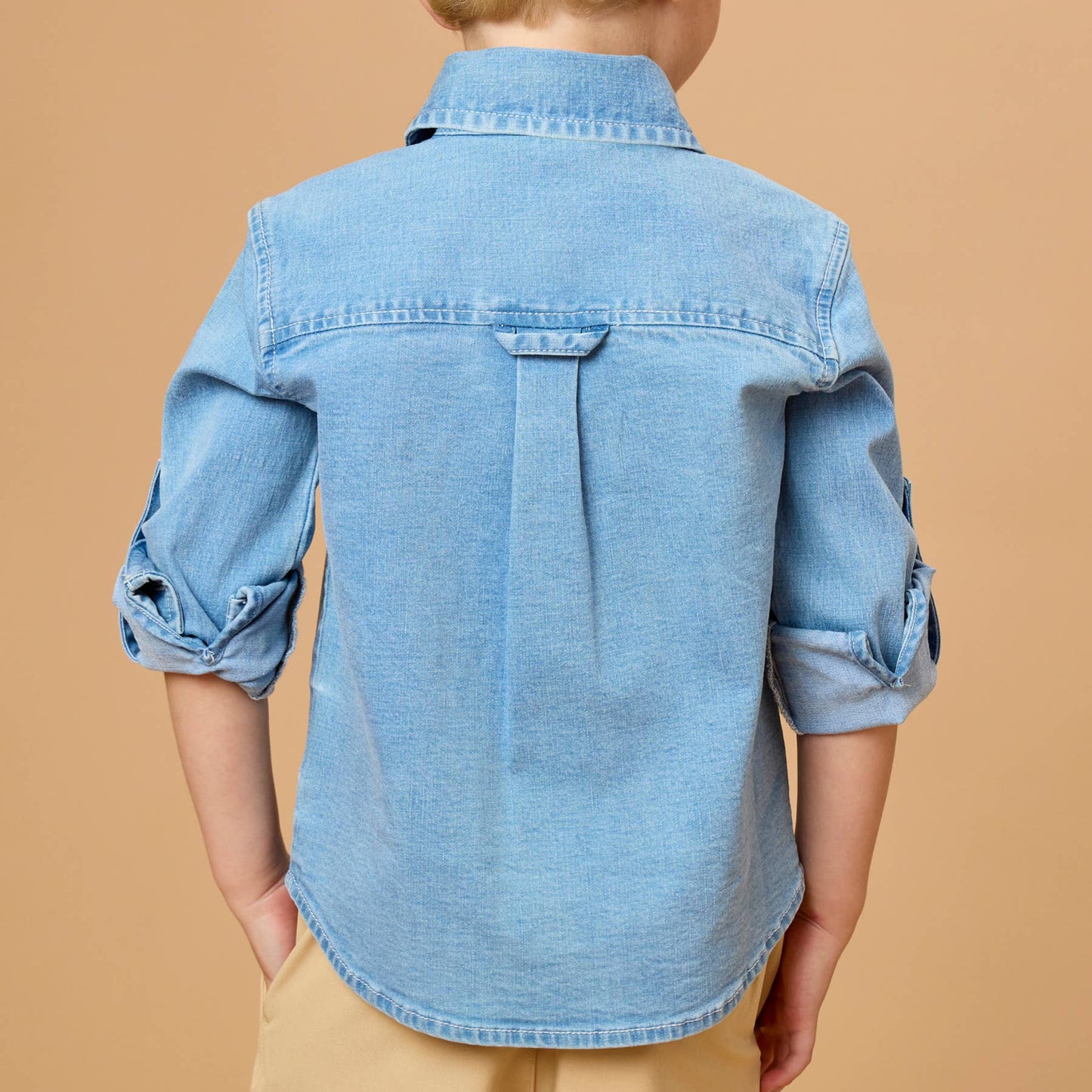 A child viewed from behind wearing a light blue denim button-up shirt with rolled sleeves and tan pants, set against a solid warm beige background.