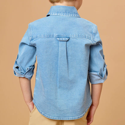 A child viewed from behind wearing a light blue denim button-up shirt with rolled sleeves and tan pants, set against a solid warm beige background.