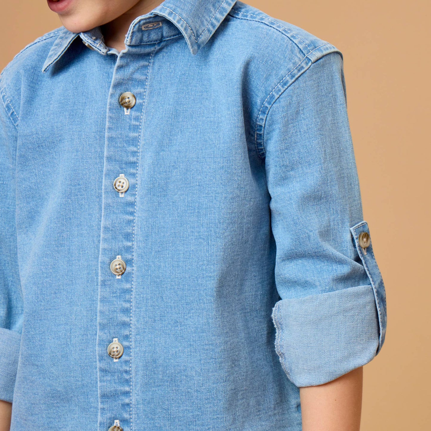 A close-up of a young boy wearing a light blue denim button-down shirt with rolled-up sleeves, set against a solid warm beige background.