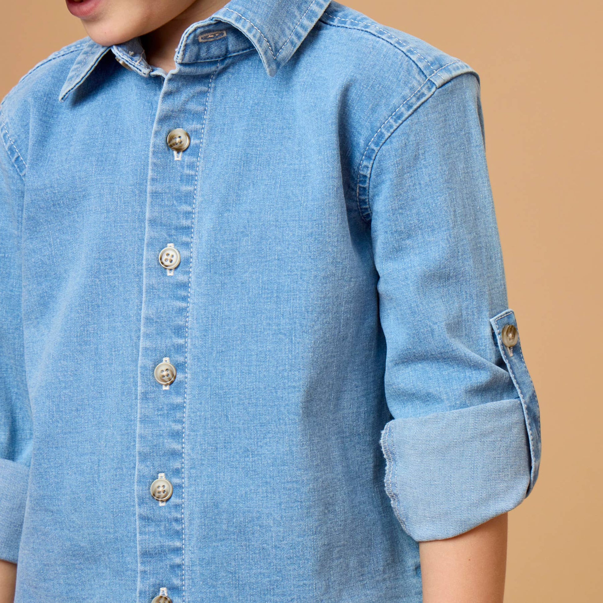 A close-up of a young boy wearing a light blue denim button-down shirt with rolled-up sleeves, set against a solid warm beige background.