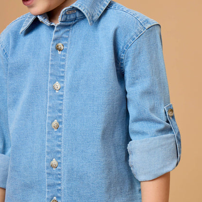 A close-up of a young boy wearing a light blue denim button-down shirt with rolled-up sleeves, set against a solid warm beige background.