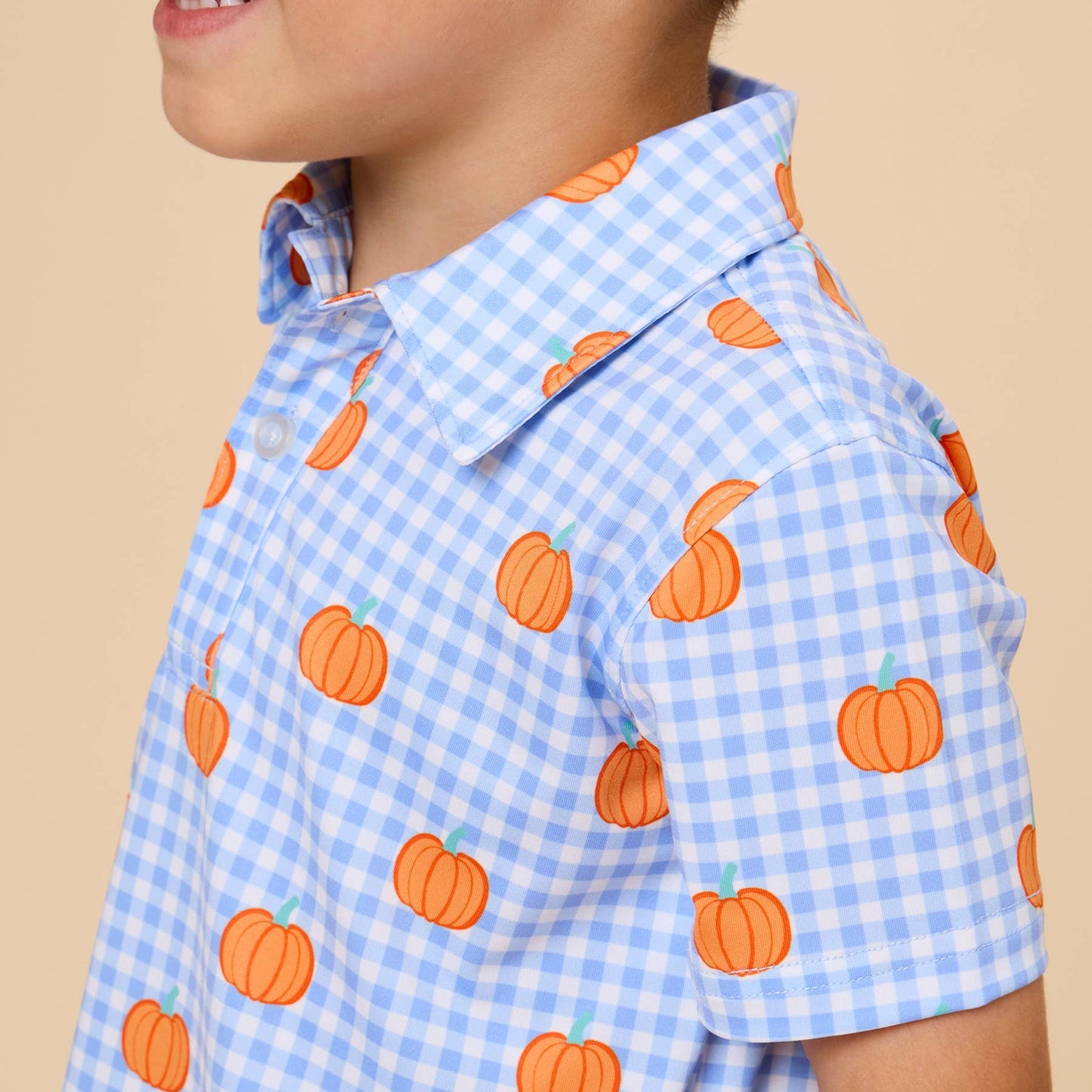 A young boy wearing a light blue and white gingham short-sleeve button-up shirt with an orange pumpkin print, shown in a side profile against a solid beige background.