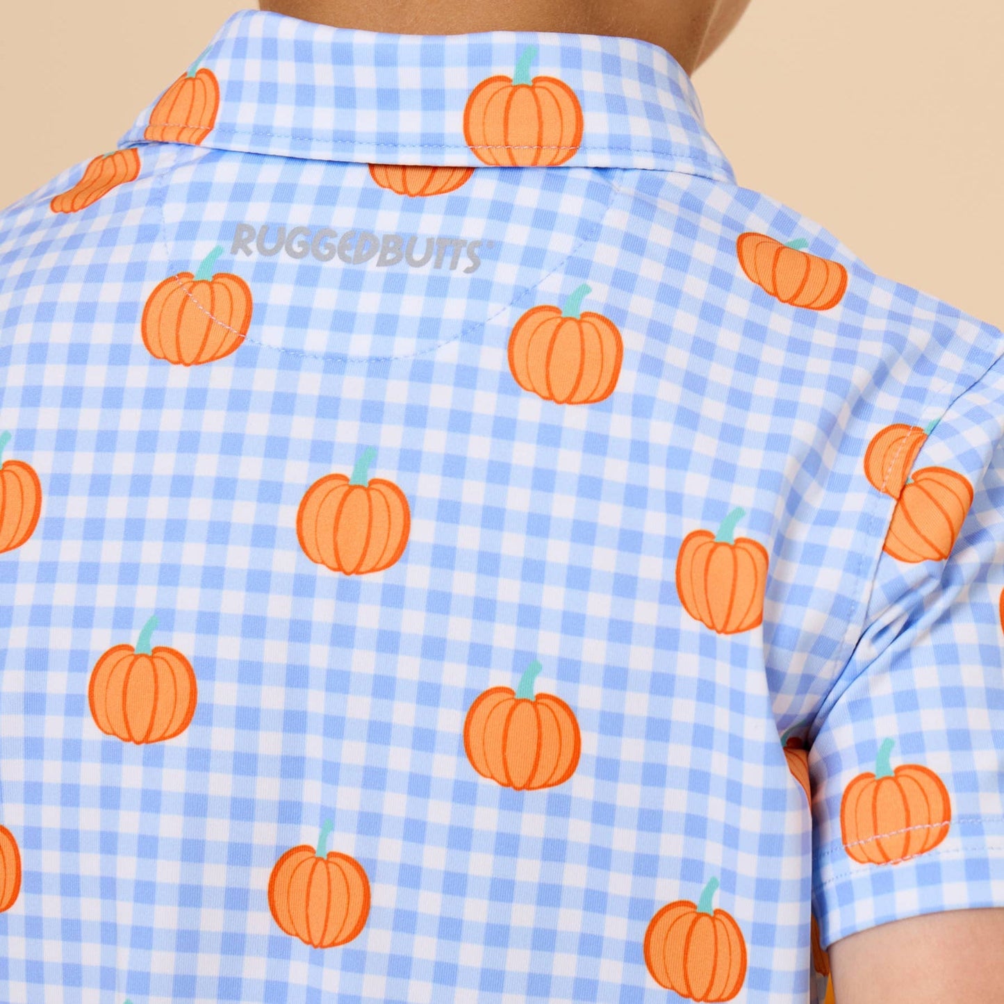 A child viewed from the back wearing a light blue and white gingham short-sleeve shirt with an orange pumpkin print. The brand name 'RUGS&BUTS' is printed in gray on the upper back.